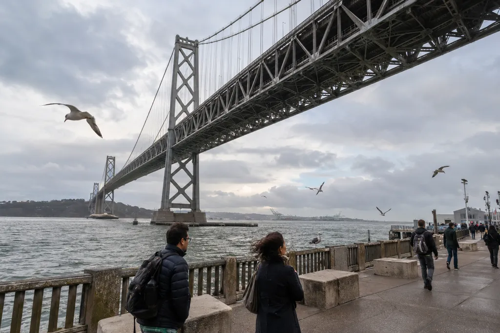 People walk along a pier under a bridge with a cloudy sky.